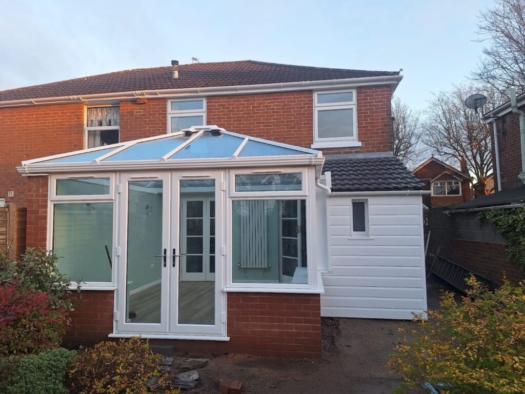 An extension to the rear of a house with a bespoke designed white UPVC conservatory with a pyramid style roof and white French Doors leading into the garden.