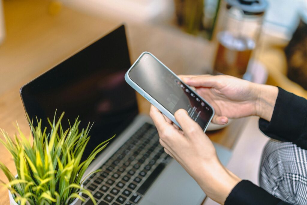 A woman holding a smartphone above a laptop in a cozy home office setting.